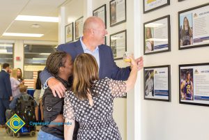 A very tall man and two women looking at framed photographs on a wall.