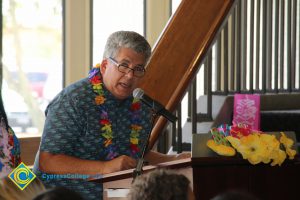 Professor Michael Brydges at microphone, wearing a lei at end-of-the-year luau
