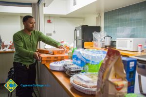 A young man in a green sweater and black pants placing pizza boxes on a counter with other food.