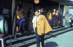 Dan Walker, the first Cypress College president walking with a gold sport coat draped over his shoulders as cheerleaders look on.