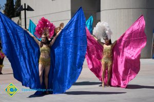 Performers wearing gold top and shorts with feathered headdress and colorful expanded fabric wings.