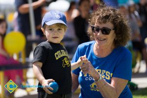 A woman in a blue Cypress College t shirt and sunglasses holding the hand of a young boy in a black t shirt, blue LA hat and blue bal.