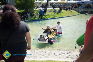 A young man wearing a black sleeveless shirt sitting in a handmade rowboat with yellow painted flames on the side and 5-gallon water jugs as wheels being pushed in the pond by someone in a white t shirt and red shorts.