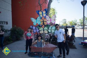 Handmade rowboat with decorative butterflies on display outside the Fine Arts building with a young man sitting inside and a young man and woman outside next to it.