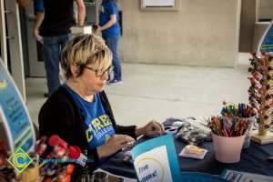 A woman with short hair and glasses and a Cypress College t shirt and black sweater sitting at an information table at the 50th Anniversary Festival and Reunion.