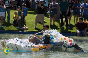 Handmade rowboat full of recyclables in the pond with spectators on the grass.