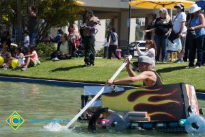 Handmade rowboat with flames painted on the sides as a young man with a black sleeveless shirt rows and people watch from the sidelines.