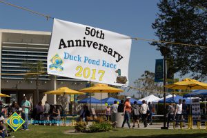 Banner hanging with words "50th Anniversary Duck Pond Race 2017" with people walking the pond area behind it.
