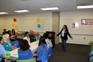 A woman in jeans, white shirt and black jacket speaks to a crowd of guests at the ESL potluck.