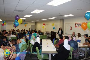 A woman in jeans, white shirt and black jacket speaks to a crowd of guests at the ESL potluck.