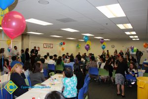 A woman in jeans, white shirt and black jacket speaks to a crowd of guests at the ESL potluck.