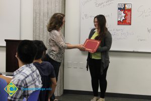 Student with long brown hair, glasses, yellow blouse and dark pants and sweater receiving an award during ESL Scholarship Potluck.