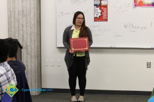 A woman wearing glasses wearing a yellow blouse and dark pants and sweater, holding a certificate.