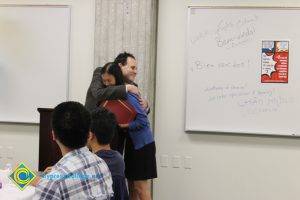 A young lady in a black dress and blue sweater receiving a hug from a gentleman during the ESL Scholarship Potluck.
