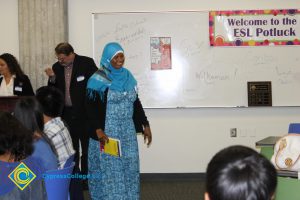 Woman in a blue and white dress and blue hijab holding a yellow book.