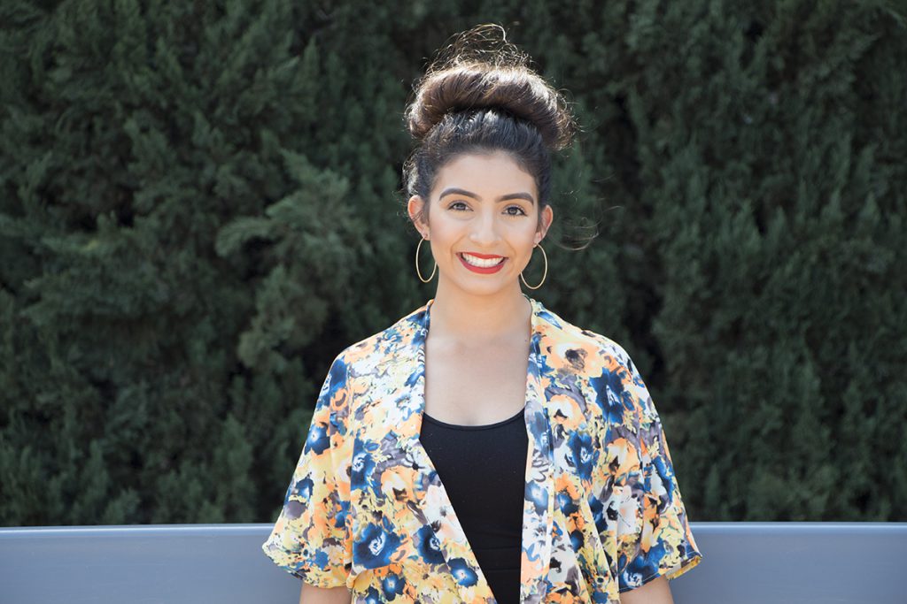 Woman with dark hair in a bun smiles, wearing hoop earrings and a flowery top