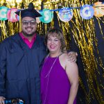 A male graduate in graduation regalia with a female in a purple dress standing in front of graduation decorations.