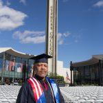 Veteran in graduation regalia standing with empty seats and the campanile in the background.