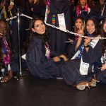 A group of five female students in graduation regalia sitting on the ground while waiting for commencement to begin.