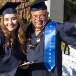 A male and female student in graduation regalia smiling.