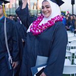 Female graduate, wearing regalia and a purple orchid lei with her hand in the air.