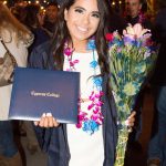 Smiling young lady in graduation regalia holding diploma and flowers.