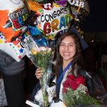 Smiling young lady in graduation regalia holding flowers and balloons.