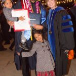 A male graduate holding a small child with another child standing in front of him, posing with Dr. Schilling.