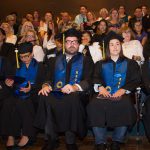 Graduates sitting in a row with guest in the background.