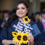 A young woman in long brown hair holding a decorated graduation cap with sunflowers.