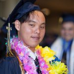 A young man wearing graduation regalia and pink and yellow leis.
