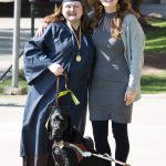 Young lady wearing graduation cap and gown with a woman in a hat and grey outfit and a black service dog,