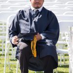 Young man wearing his graduation gown and holding his cap and tassel, sitting in a chair with empty white chairs in the background,