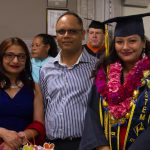 Young STEM student in her graduation regalia and floral leis with a man and woman by her side and various staff in the background.