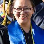 A young lady with glasses and wearing her cap and gown, smiling during commencement.