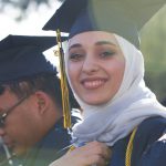 A smiling young lady in her cap and gown standing next to a young man in his cap and gown who is looking downward.