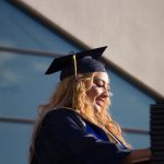 Young lady with long blond hair in graduation cap and gown, speaking at the podium.