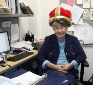 A woman sits in an office while wearing a red velvet hat with gold trim