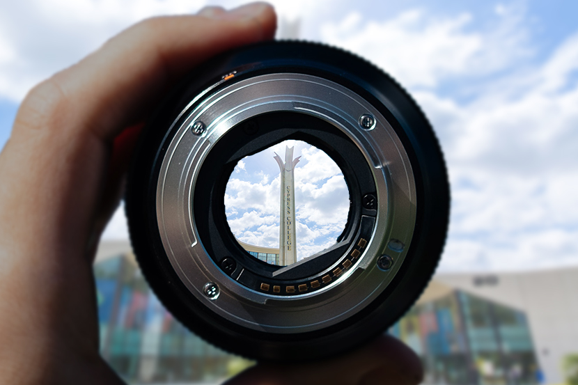 Hand holding camera lens with the focus on the campanile