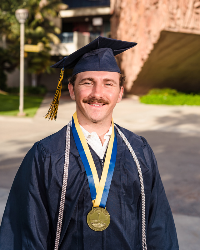 Student Garrett Deiro stands in front of a Cypress College building while wearing graduation regalia.