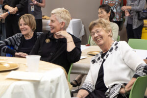 Three woman sit at a table in a crowded room. They are laughing.