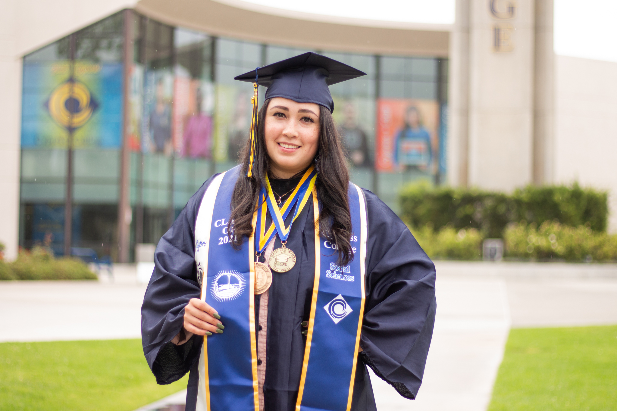 Student Geraldine Lopez in graduation regalia stands in front of the Cypress College Student Life and Leadership building.