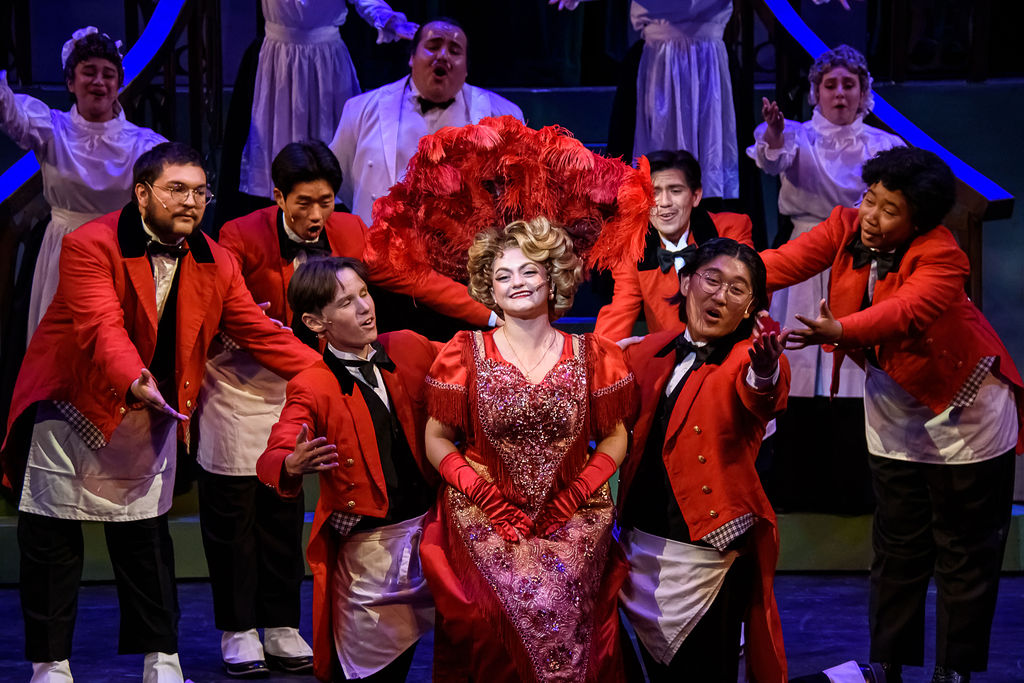 The lead of the musical "Hello, Dolly!" sits on the knees of actors dressed as waitstaff in this turn-of-the-century-set performance.