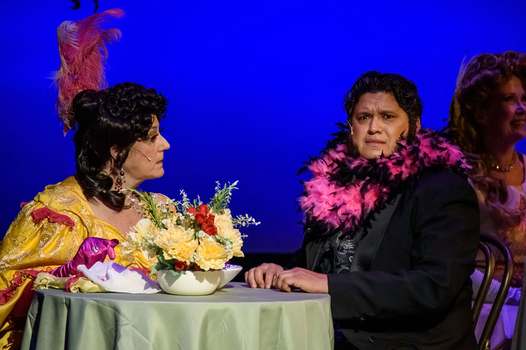 A couple in 1890s costumes sit at a cafe table in a scene from "Hello, Dolly!"
