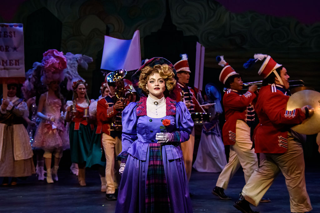 Woman in purple satin 1890s costume holds a note in front of a marching band in the musical "Hello, Dolly!"