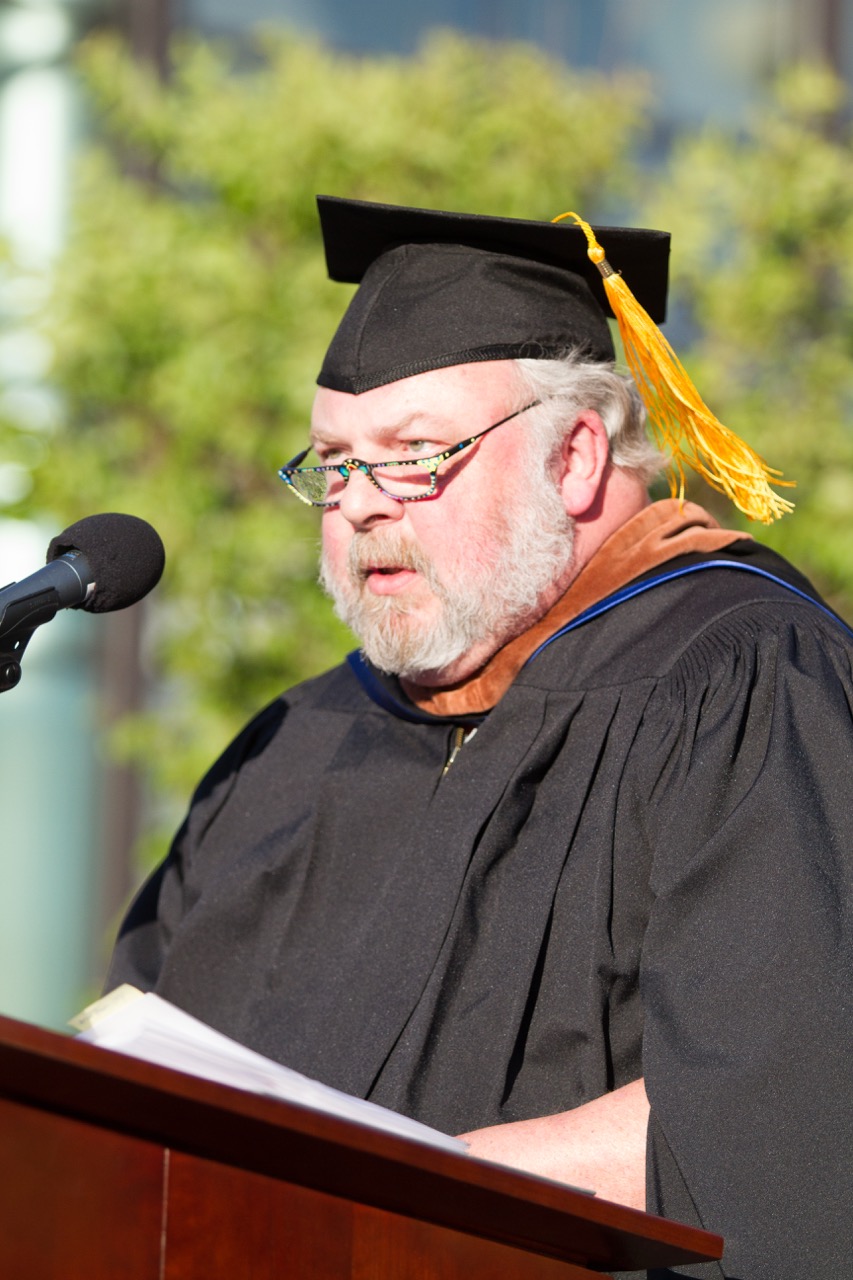 A man wearing academic regalia and glasses is speaking at a podium with a microphone outdoors.