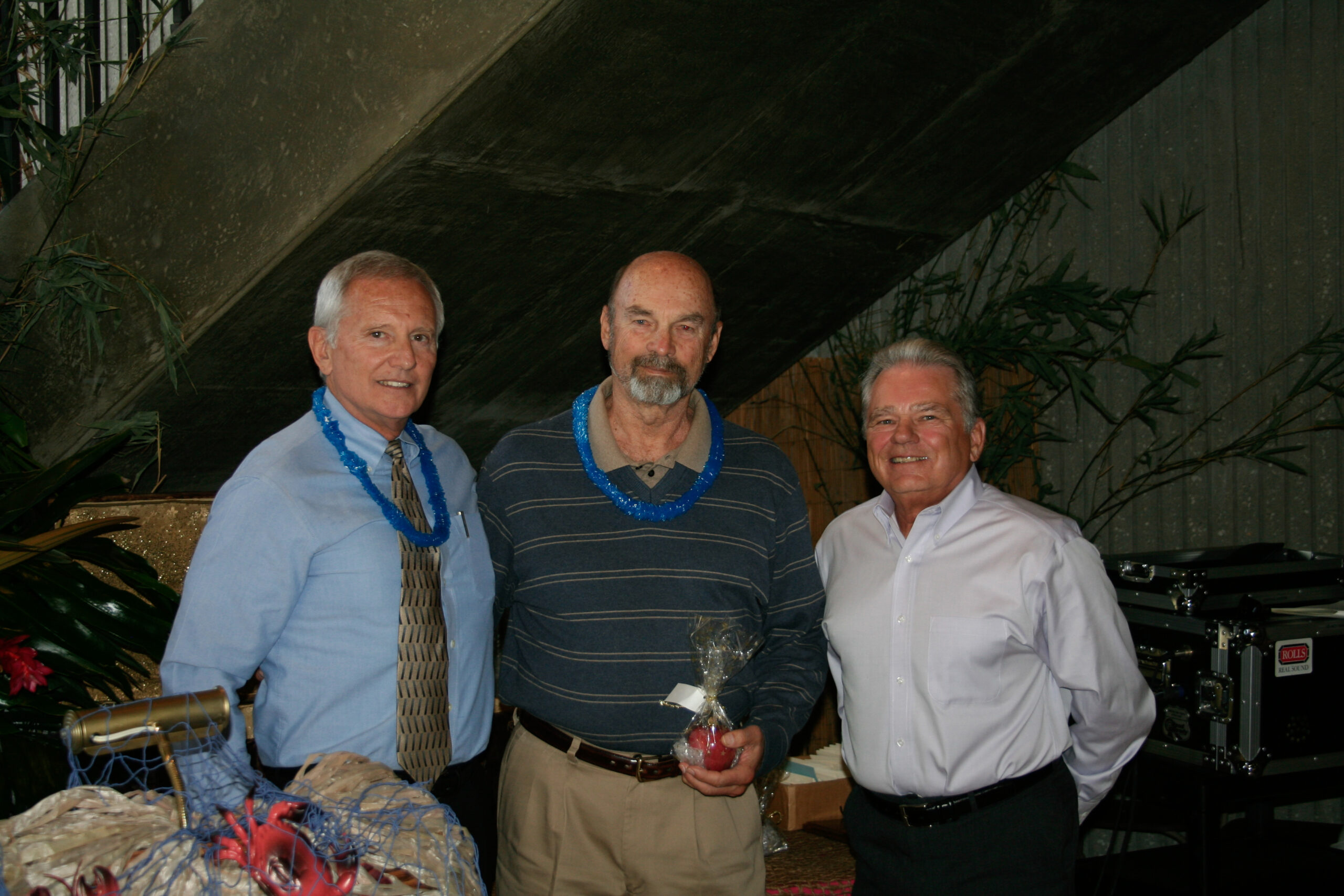 Three men pose for a photograph at a ceremony. The man in the middle is holding an award that is shaped like an apple.