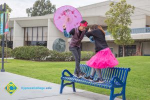 Young man with umbrella and young lady with pink tutu dancing on a blue bench.