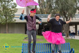 Young man with umbrella and young lady with pink tutu dancing on a blue bench.