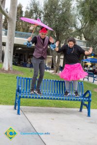 Young man with umbrella and young lady with pink tutu dancing on a blue bench.